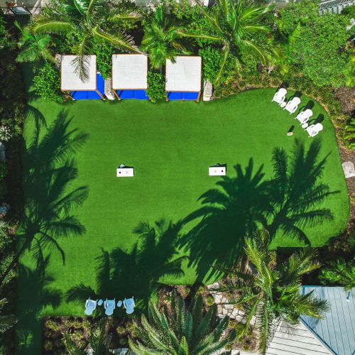 The image shows a grassy area with lawn games, surrounded by trees and foliage. There are three cabanas, several lounge chairs, and shadows of palm trees.