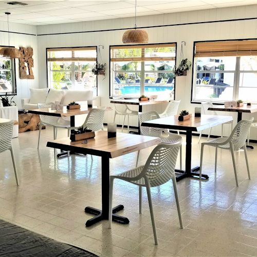 The image shows a bright, airy café with white furniture, several tables and chairs, large windows, and light streaming in from outside.