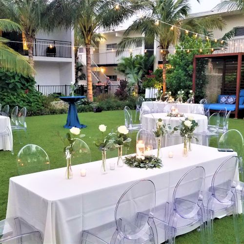 An outdoor event setup with white-clothed tables, transparent chairs, flowers in vases, string lights, and a greenery backdrop surrounded by buildings.