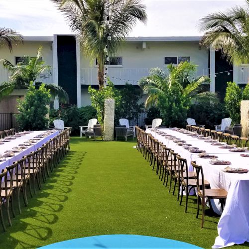 An outdoor setting with two long tables covered with white cloth, surrounded by chairs, set for a meal in front of a building and palm trees.