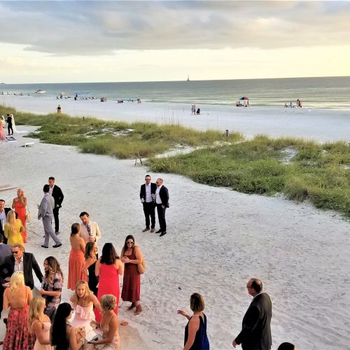 A group of people are gathered on a beach, wearing formal attire and socializing as the sun sets in the background, with the sea and sky visible.