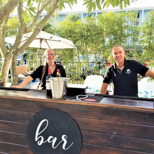 Two people standing behind an outdoor bar with a tree and greenery in the background. The bar has a "bar" sign on it.