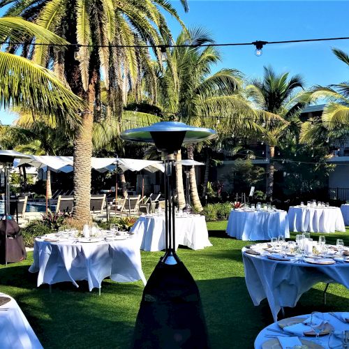 An outdoor venue with palm trees features round tables set for a meal, string lights overhead, and patio heaters scattered throughout the area.