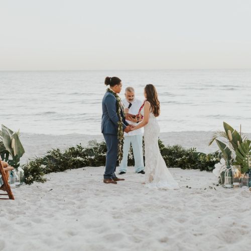 A couple is getting married on a beach, surrounded by close friends and family, with the ocean in the background, during a beautifully serene ceremony.