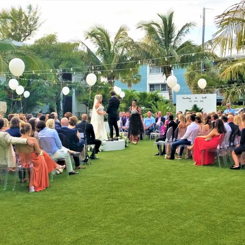 A wedding ceremony is taking place outdoors with guests seated on either side of the aisle, under string lights and white balloons, surrounded by greenery.