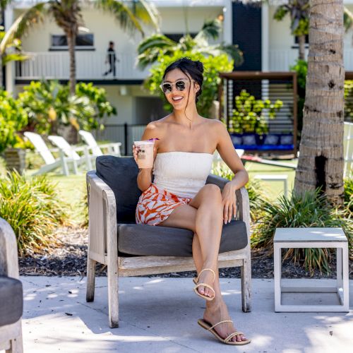 A woman wearing sunglasses sits on an outdoor chair holding a drink, surrounded by palm trees and resort-style furniture.