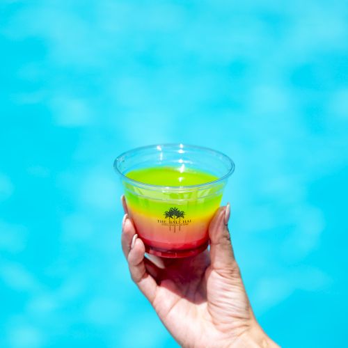 A hand holds a multi-colored drink in a clear plastic cup against a bright blue watery background.