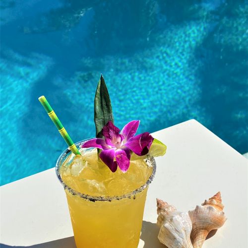 A tropical drink garnished with a flower and a lime slice sits on a poolside table next to two seashells with a blue pool in the background.