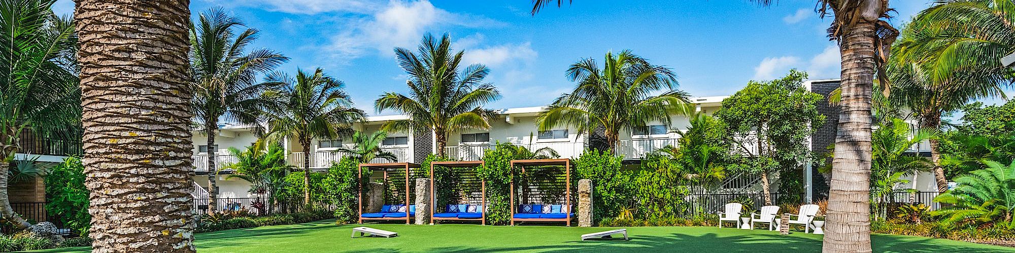 A lush resort-like garden features palm trees, green grass, shaded seating areas, and a stone pathway under a bright blue sky ending the sentence.