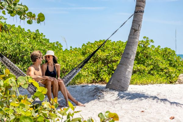 A couple is sitting together in a hammock tied between trees on a sandy beach, surrounded by lush greenery, under a clear blue sky.