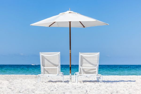 Two white lounge chairs and an umbrella on a sandy beach facing a calm blue sea under a clear sky.