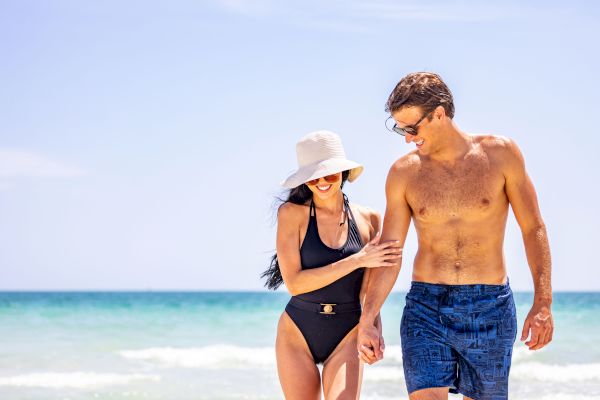 Two people, a woman in a black swimsuit and sunhat, and a shirtless man in swim trunks, walk on a beach near the ocean, smiling together.