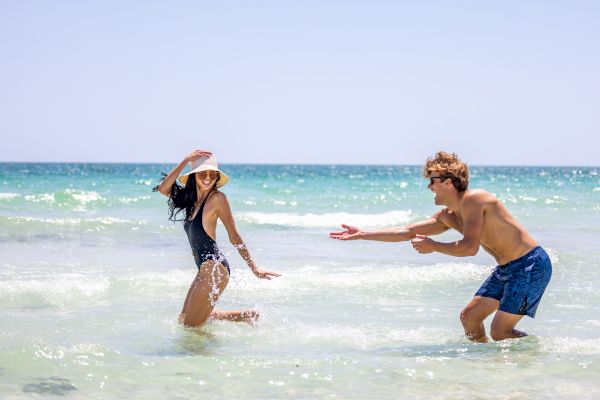 A man and woman are playfully splashing water on a sunny beach, with clear blue skies and waves in the background.