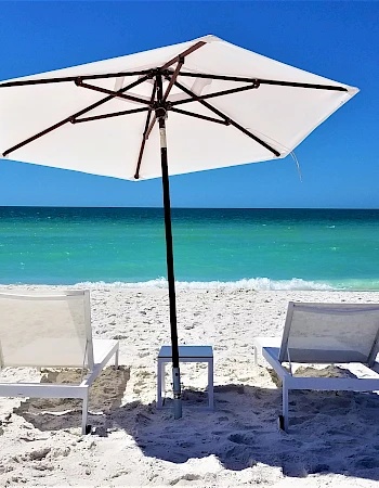 Two lounge chairs and a white umbrella on a sandy beach with clear blue water and sky in the background. Cozy seaside relaxation spot.