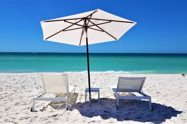 Two lounge chairs and a white umbrella on a sandy beach facing clear blue water under a bright sky.